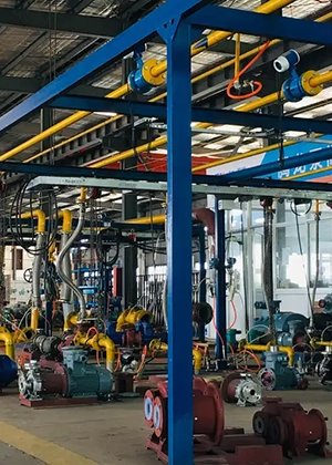 Industrial workshop interior with various machinery, pipes, and metal frames. Multiple cables and tubes hang from the ceiling, and various colored machines are placed on the floor.